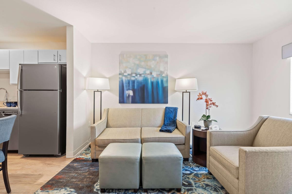 A modern living room with a sofa, ottoman, and two side tables with lamps. A kitchen area with a refrigerator is visible in the background.