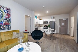A modern apartment with a yellow sofa, a dining table with white chairs, a kitchen with white cabinets, and a colorful abstract painting on the wall.