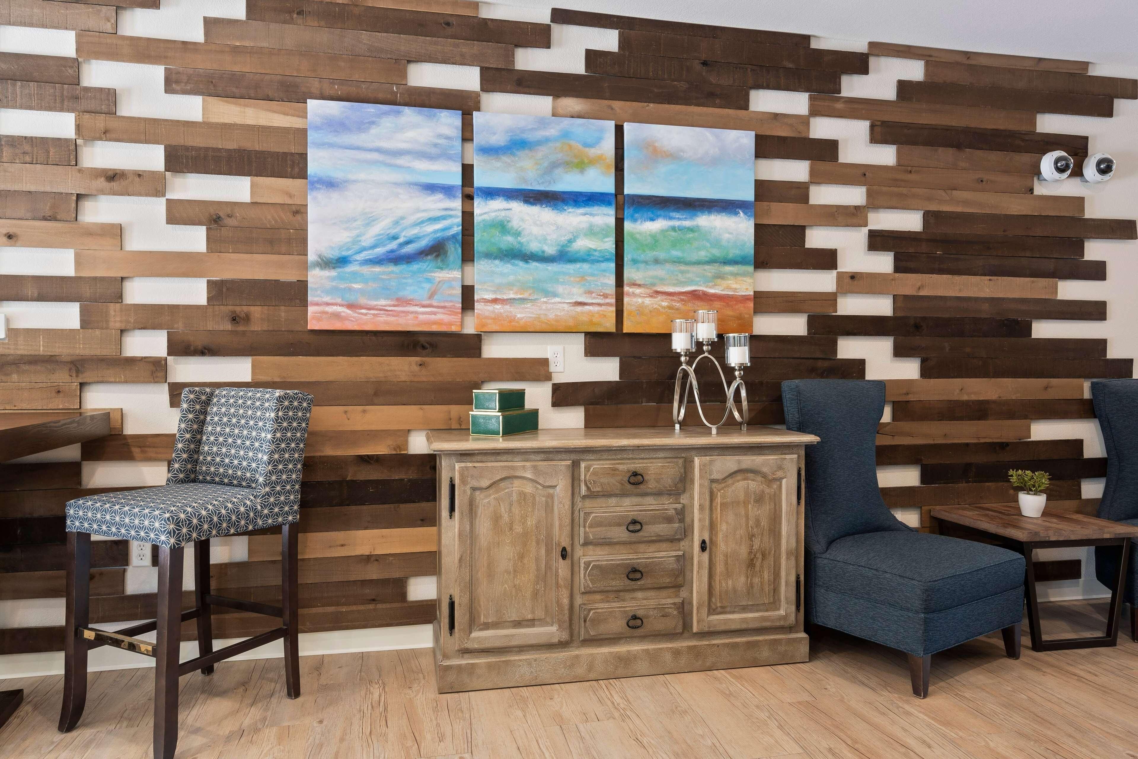Una habitación con panel de madera, un armario de madera con cajones, un sillón azul, una silla estampada y una pequeña mesa con una planta.