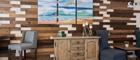 Una habitación con panel de madera, un armario de madera con cajones, un sillón azul, una silla estampada y una pequeña mesa con una planta.