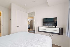 A bedroom with a bed, a TV mounted on a wall, and an open door leading to a kitchen. - Waterwalk Extended Stay Kansas City - Overland Pk (Overland Park)