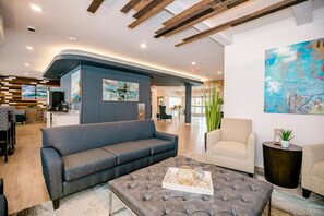 A modern lobby with a grey sofa, a tufted ottoman, and a coffee table. There is a reception desk with a blue accent wall and artwork. - Waterwalk Extended Stay Kansas City - Overland Pk (Overland Park)