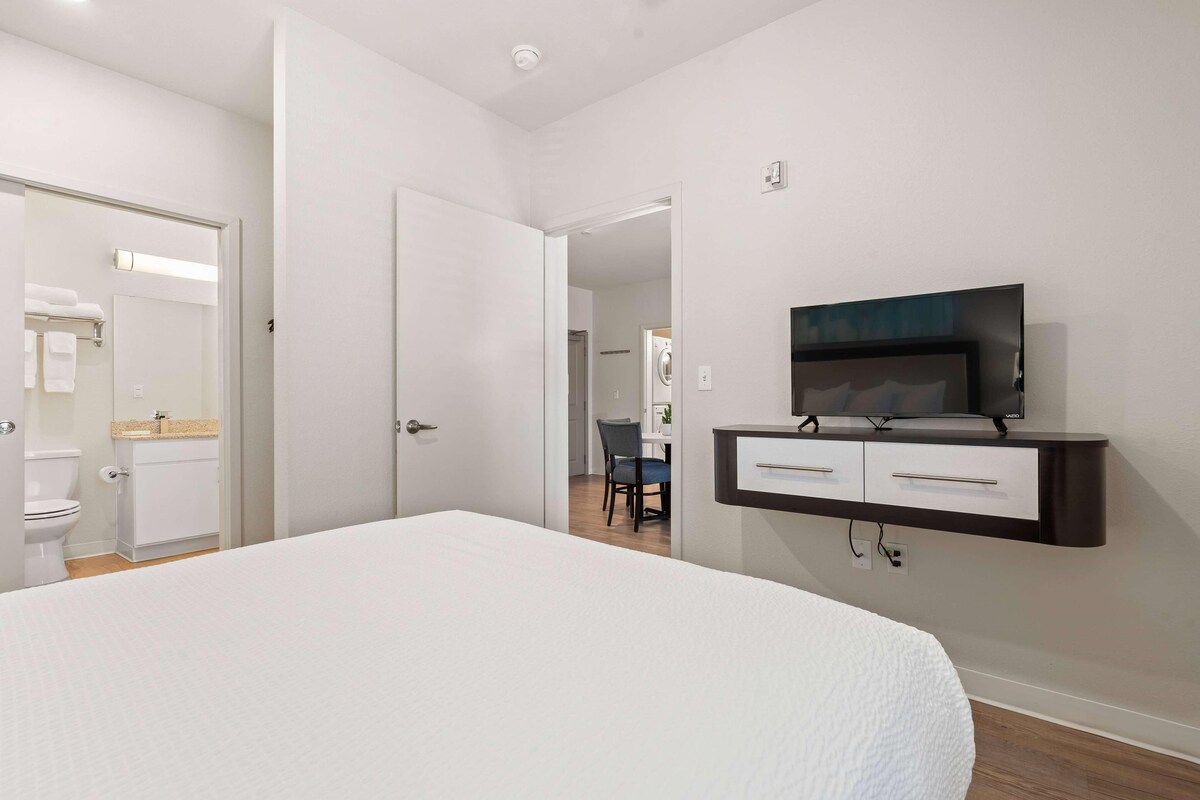 Une chambre à coucher avec un grand lit, un téléviseur fixé au mur et une salle de bain visible à travers une porte ouverte.