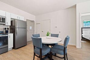 A modern kitchen with stainless steel appliances, a dining area with a round table and blue chairs, and a white door with a small plant on it. - Waterwalk Extended Stay Kansas City - Overland Pk (Overland Park)
