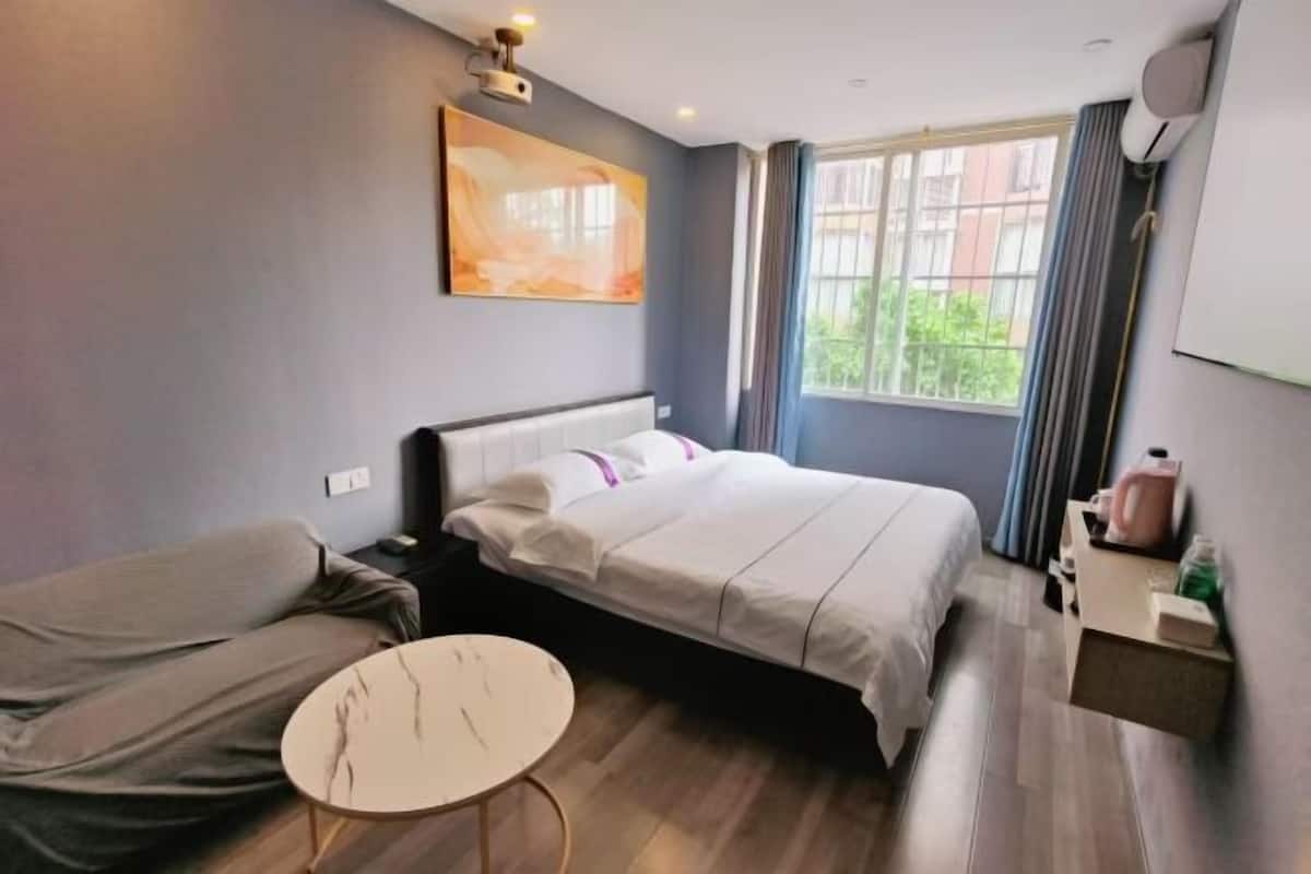 Habitación de hotel con cama, una mesa redonda, un cuadro en la pared, una ventana con cortinas y una mesita de noche con lámpara.