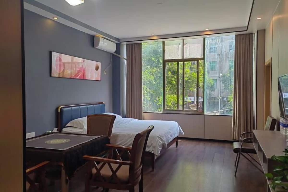 Habitación de hotel con cama, escritorio, silla, televisor y ventana con vista a follaje.