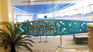 Et receptionsområde med et skilt der viser 'PISCINE', en potteplante og et glimt af et poolområde.