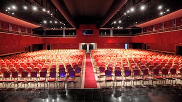 Een ruime zaal met rijen rode en paarse stoelen, een middenpad en een rood tapijt.