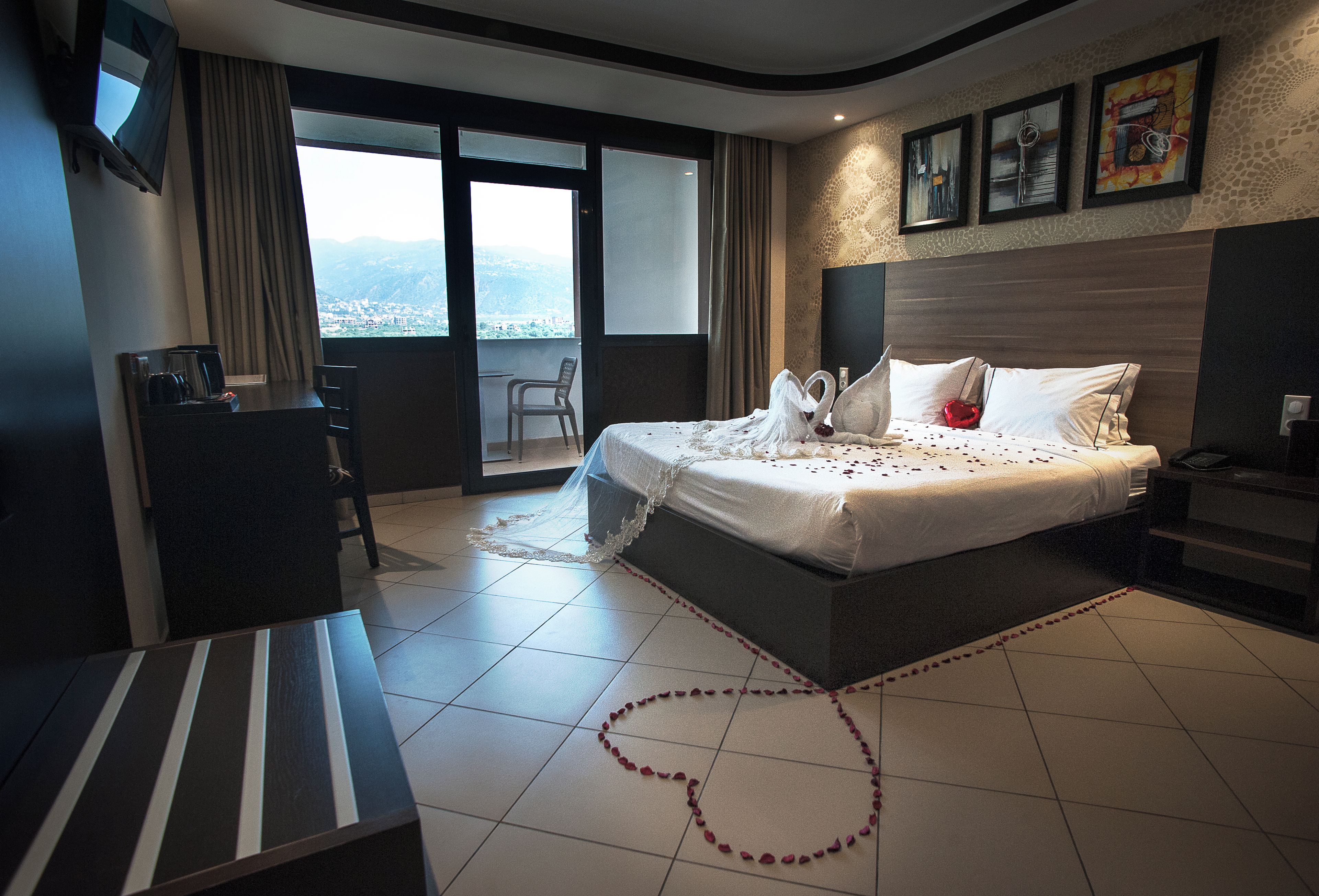 A hotel room with a large bed, a desk, a chair, and a balcony with a city view.
