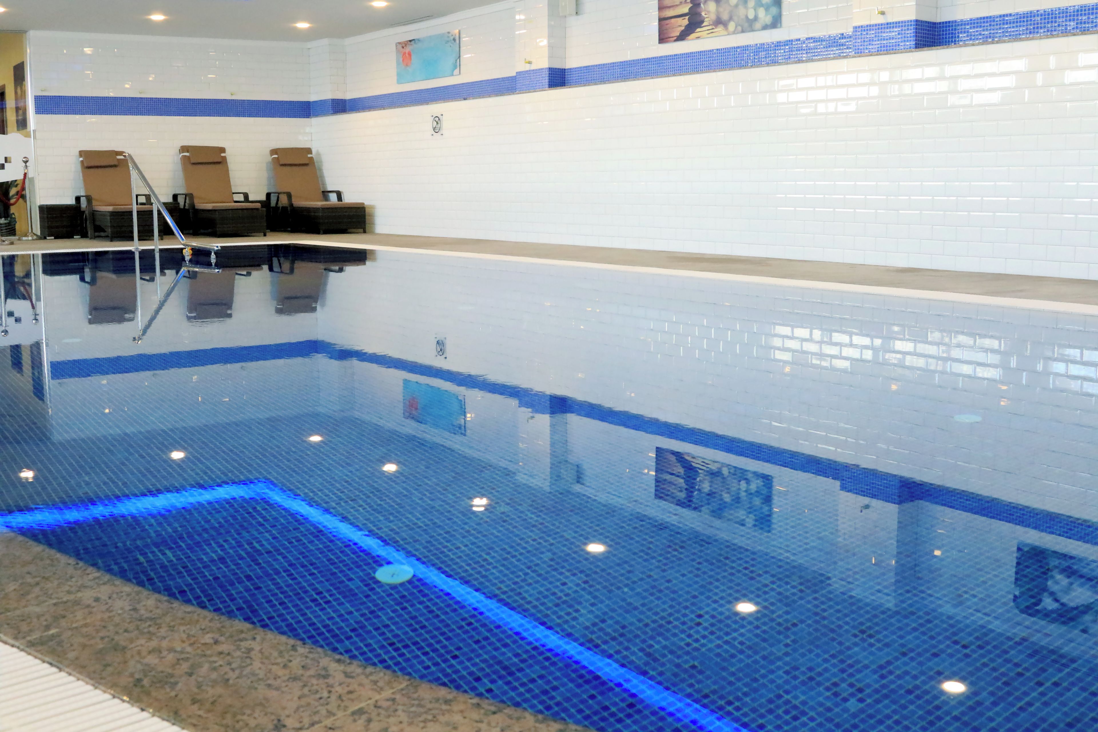 Piscina com água azul cristalina, parede azulejada e três espreguiçadeiras.