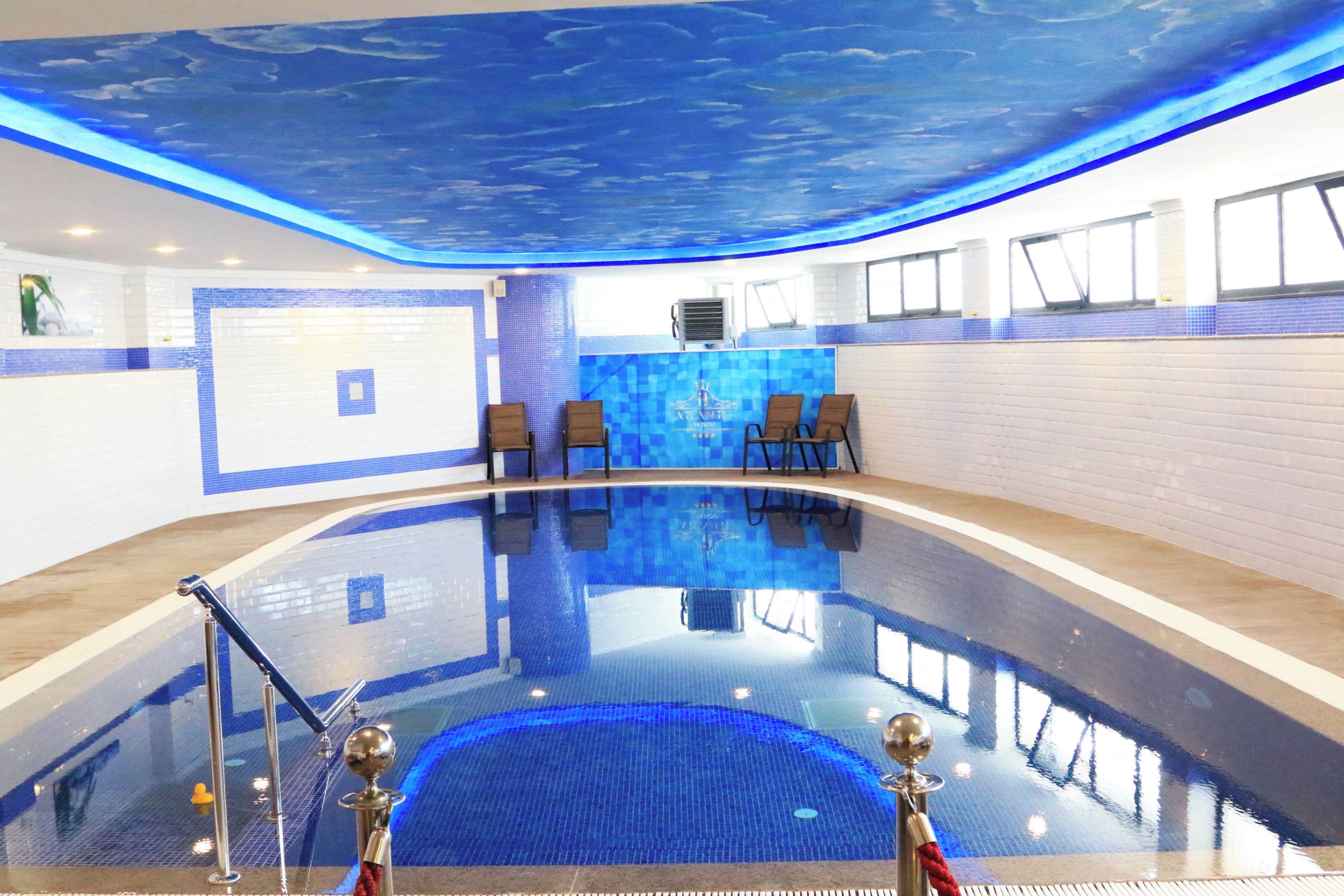 A swimming pool with a blue ceiling, lounge chairs, and a tiled wall.