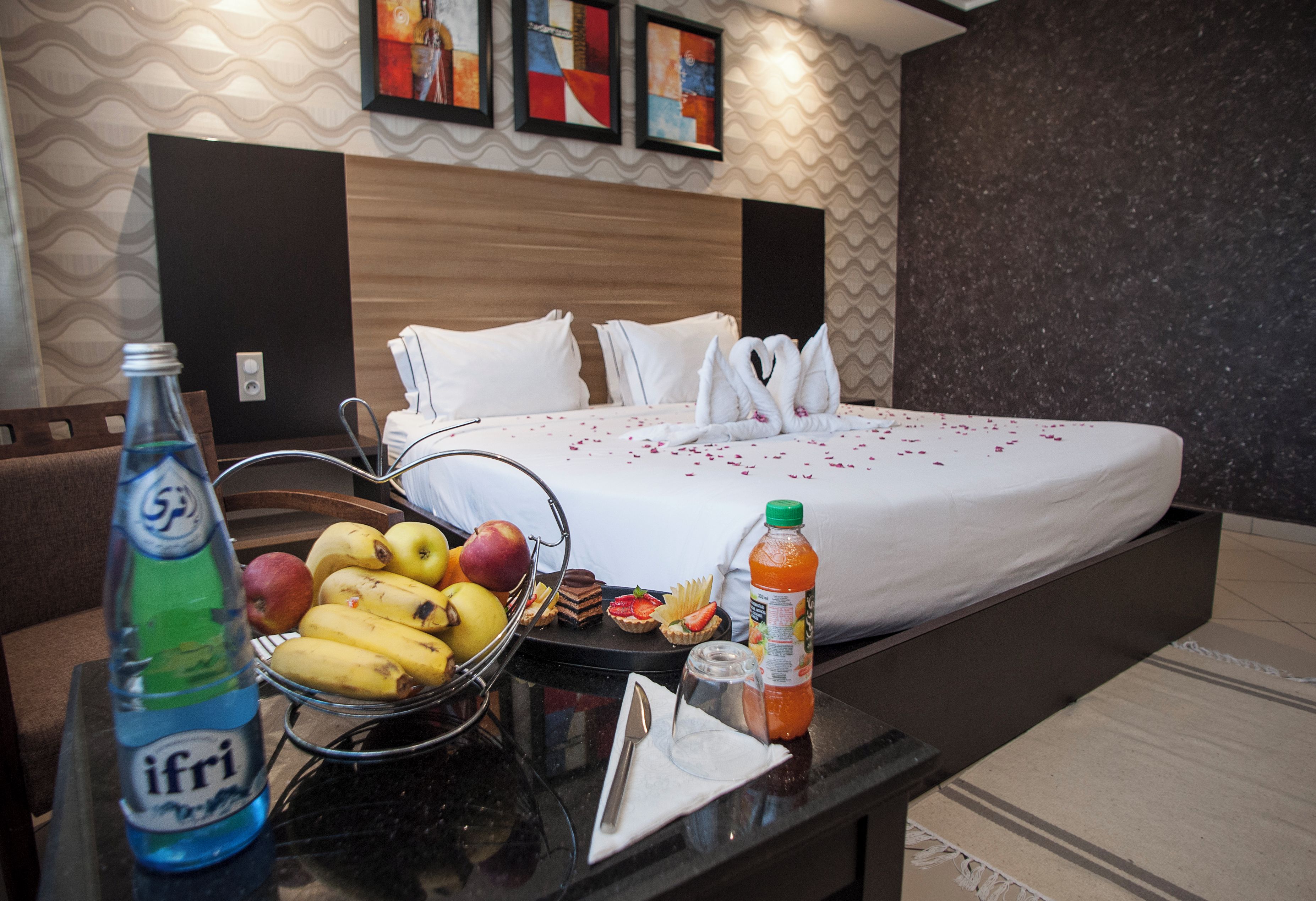 A hotel room with a bed, a dining table with fruits and a bottle, and a wall with artwork.
