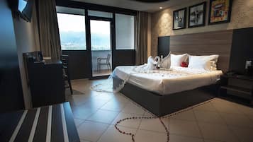 A hotel room with a large bed, a desk, a chair, and a balcony with a city view.