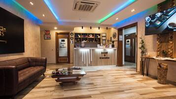 A hotel reception area with a reception desk, a brown leather sofa, a coffee table, and a television mounted on the wall.