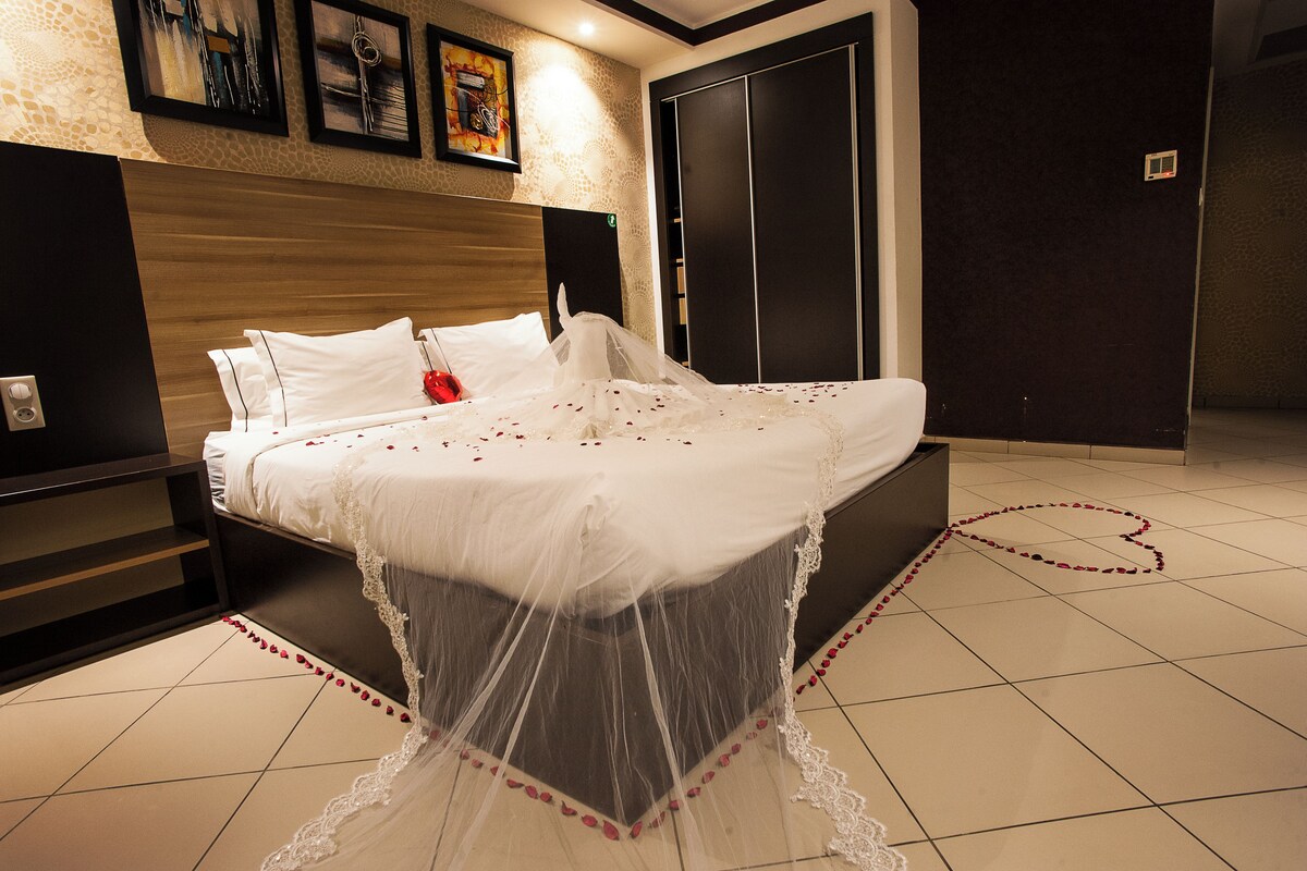 A neatly made bed with a canopy, a bedside table, and a wall-mounted power outlet.