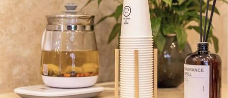 A tea set with a glass container and a stack of white paper cups, a bottle of fragrance oil, and a potted plant.