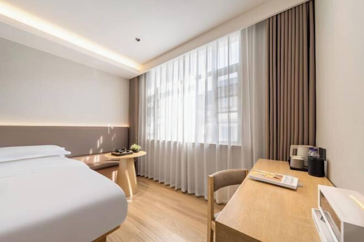 A hotel room with a bed, a desk, a chair, a small table, and a window with curtains.