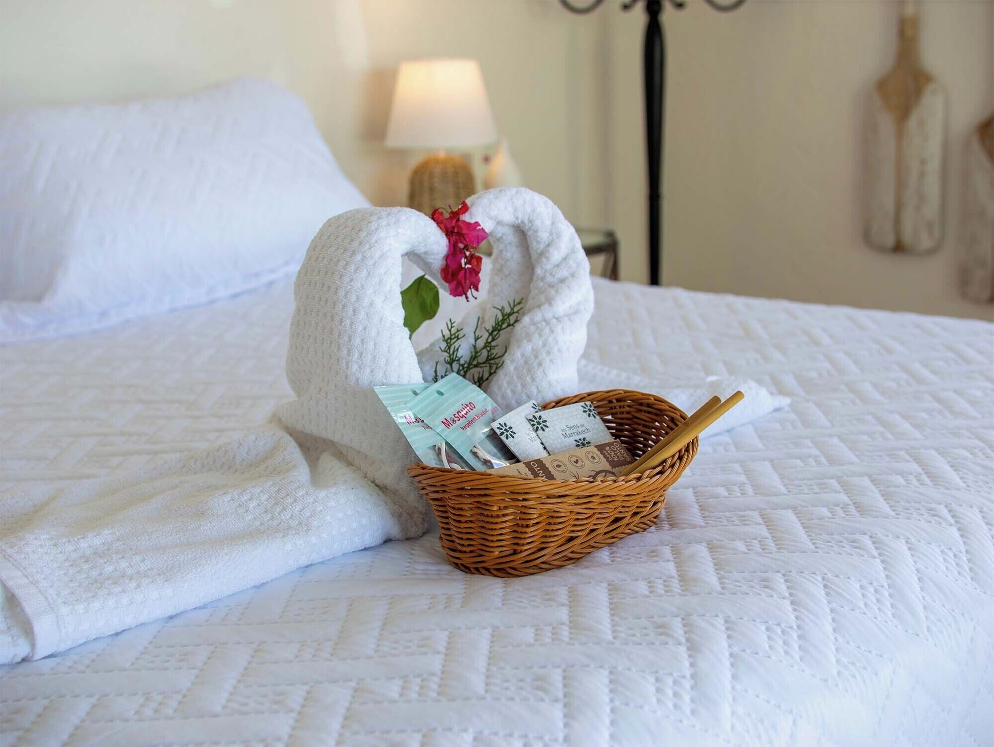 A neatly made bed with a white quilt, a white towel shaped into a heart, and a wicker basket containing a tissue box and a small card.
