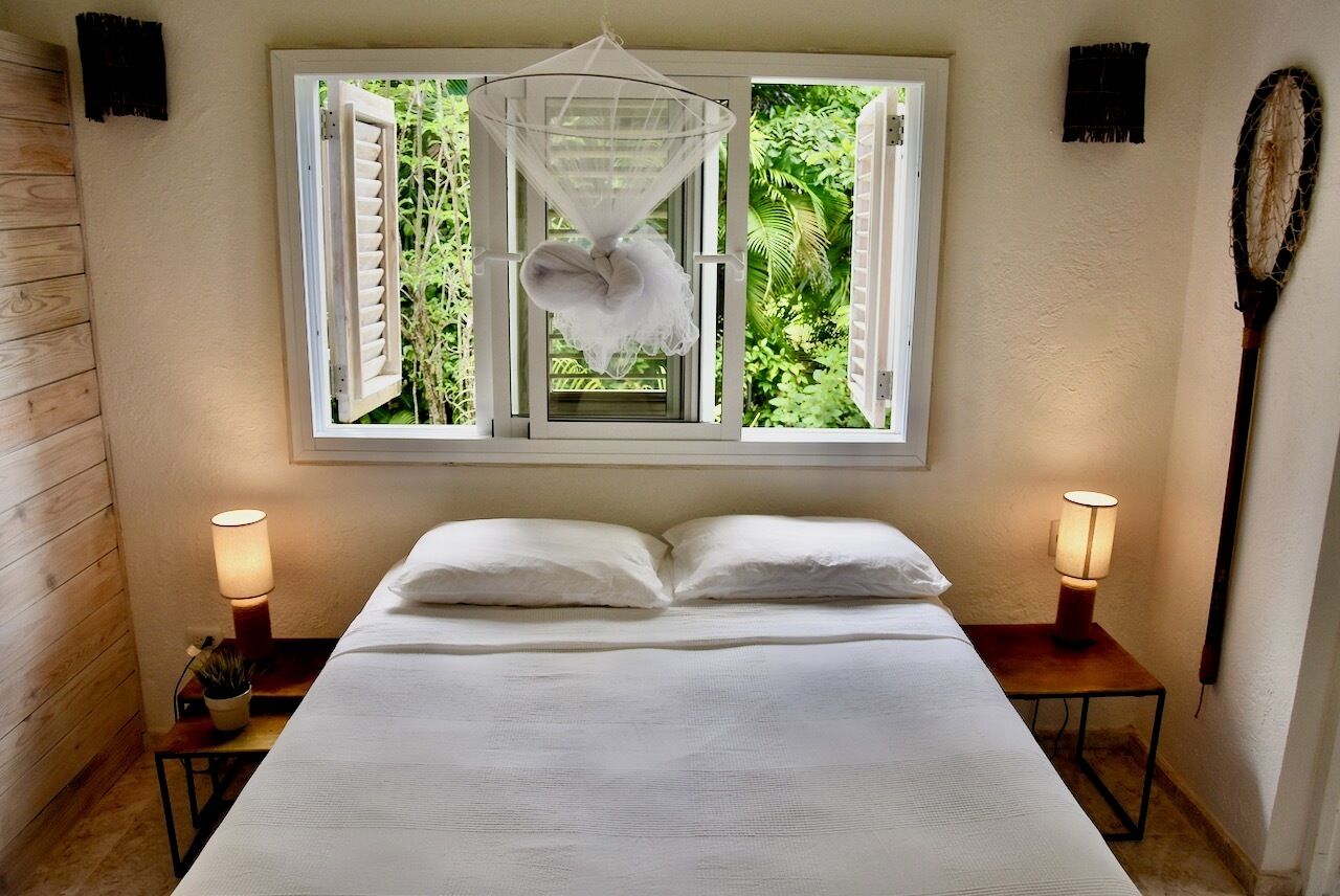A bedroom with a bed, two bedside lamps, a window with a view of greenery, and a hanging net.