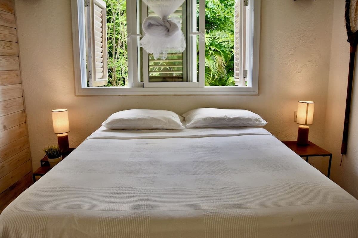 A neatly made bed with white linens, two bedside lamps, and a window with a view of greenery.