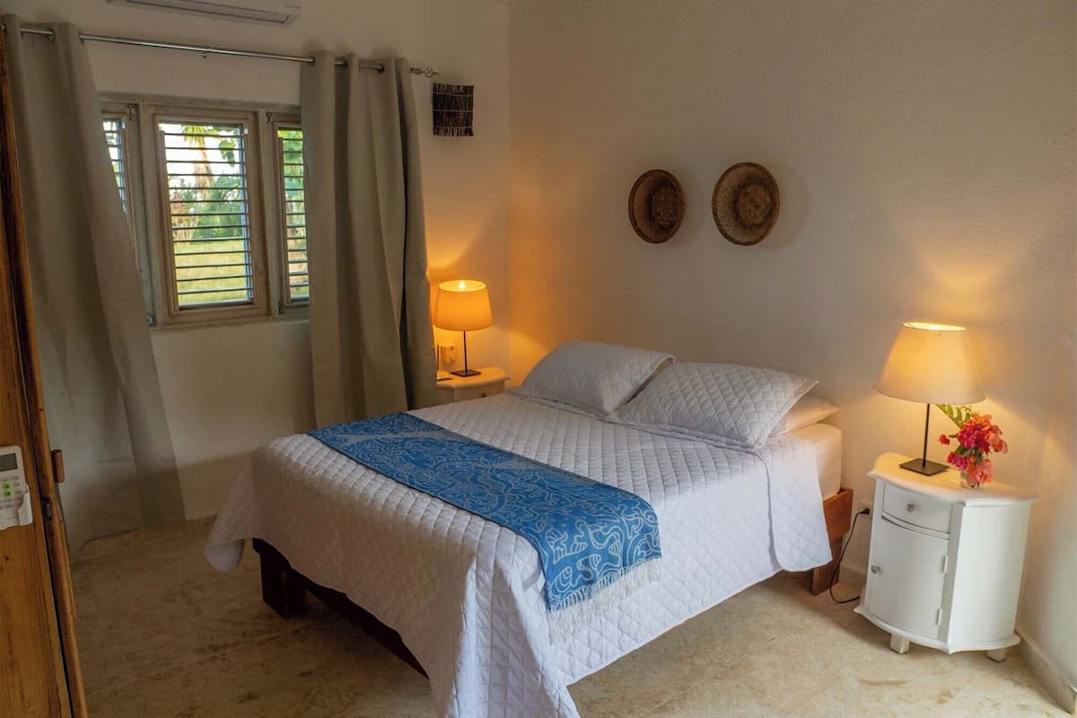A bedroom with a bed, two bedside tables, a nightstand, a window with curtains, and a wall-mounted light fixture.
