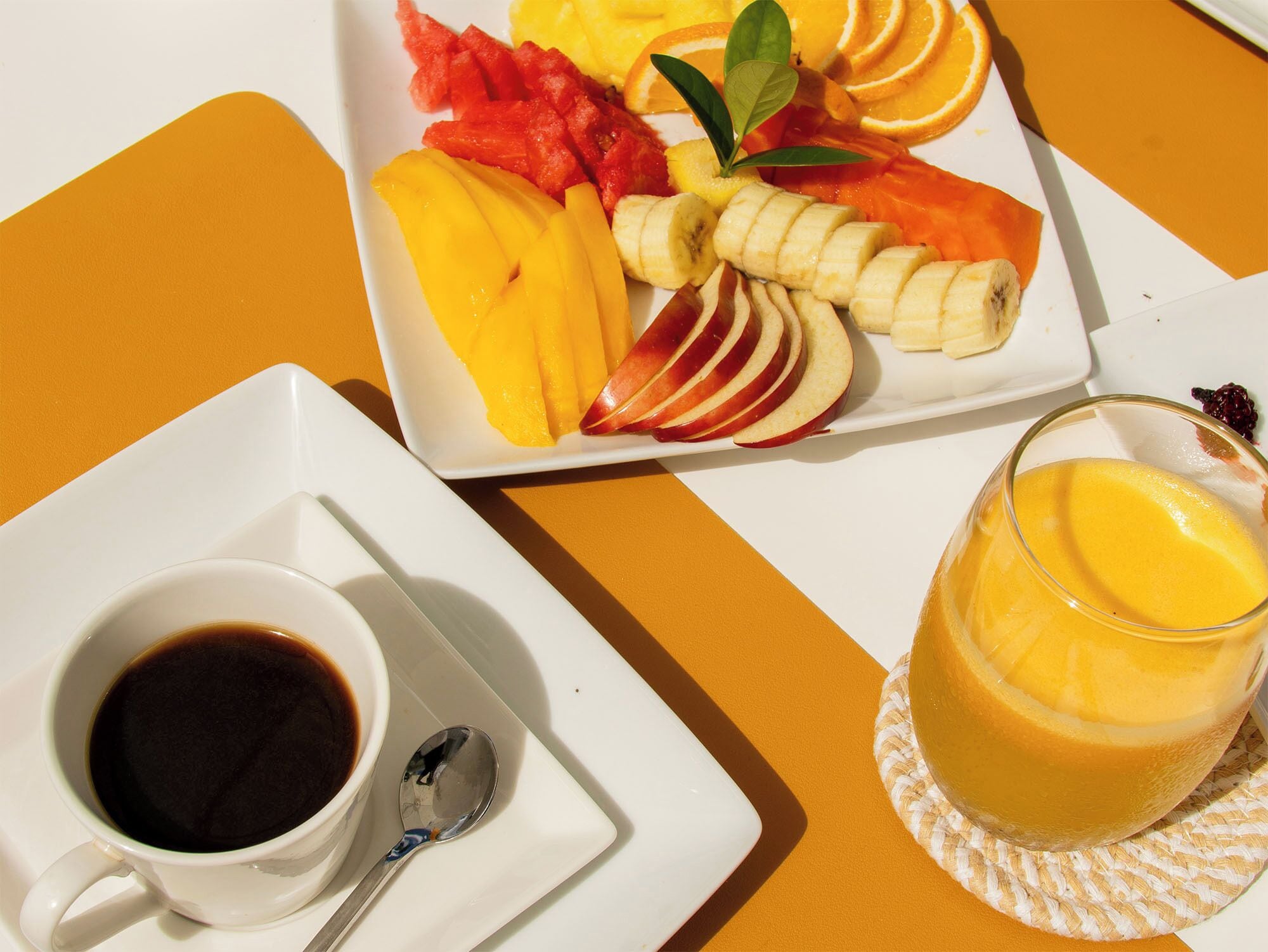 Une table mise pour le petit-déjeuner, avec une assiette de fruits coupés et un verre de jus d’orange.