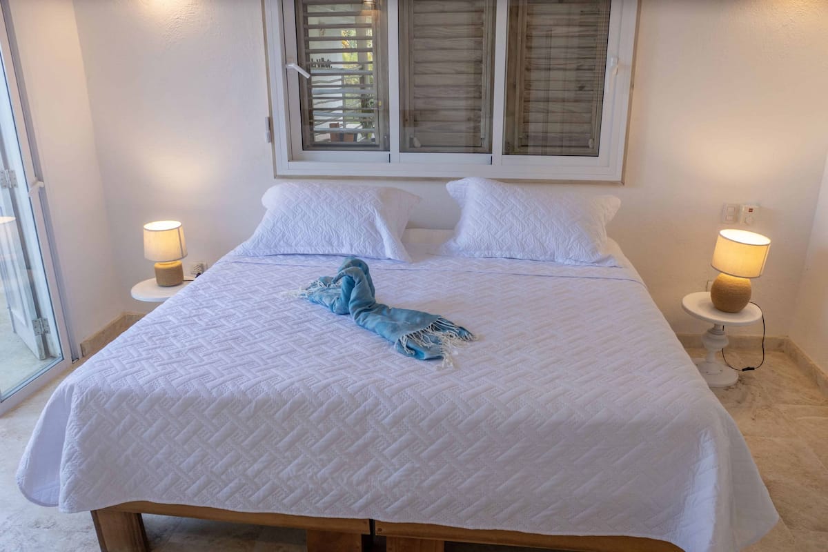 A neatly made bed with a quilted white bedspread, two white pillows, and a blue towel on the bed. There are two bedside lamps on white stands, and a window with blinds above the bed.