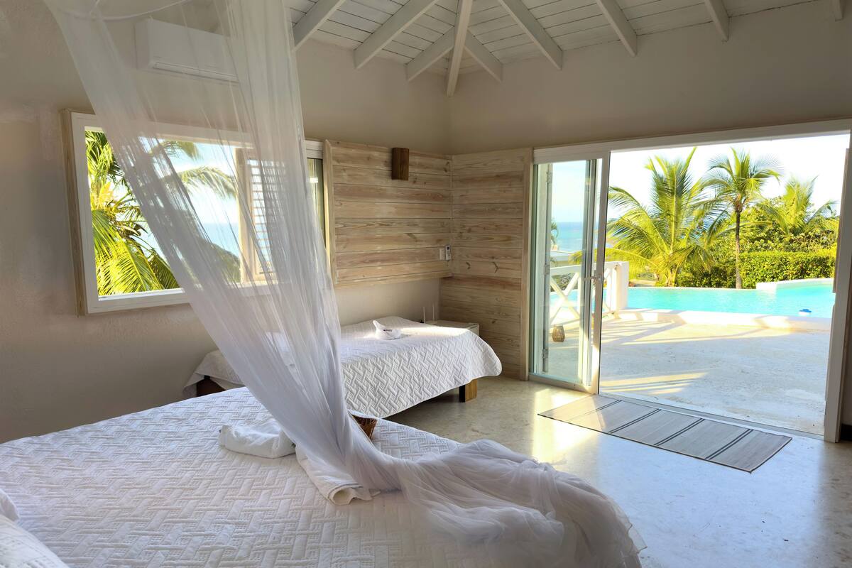 A bedroom with two beds, a large window with a view of the ocean, and a sliding glass door leading to a pool area.