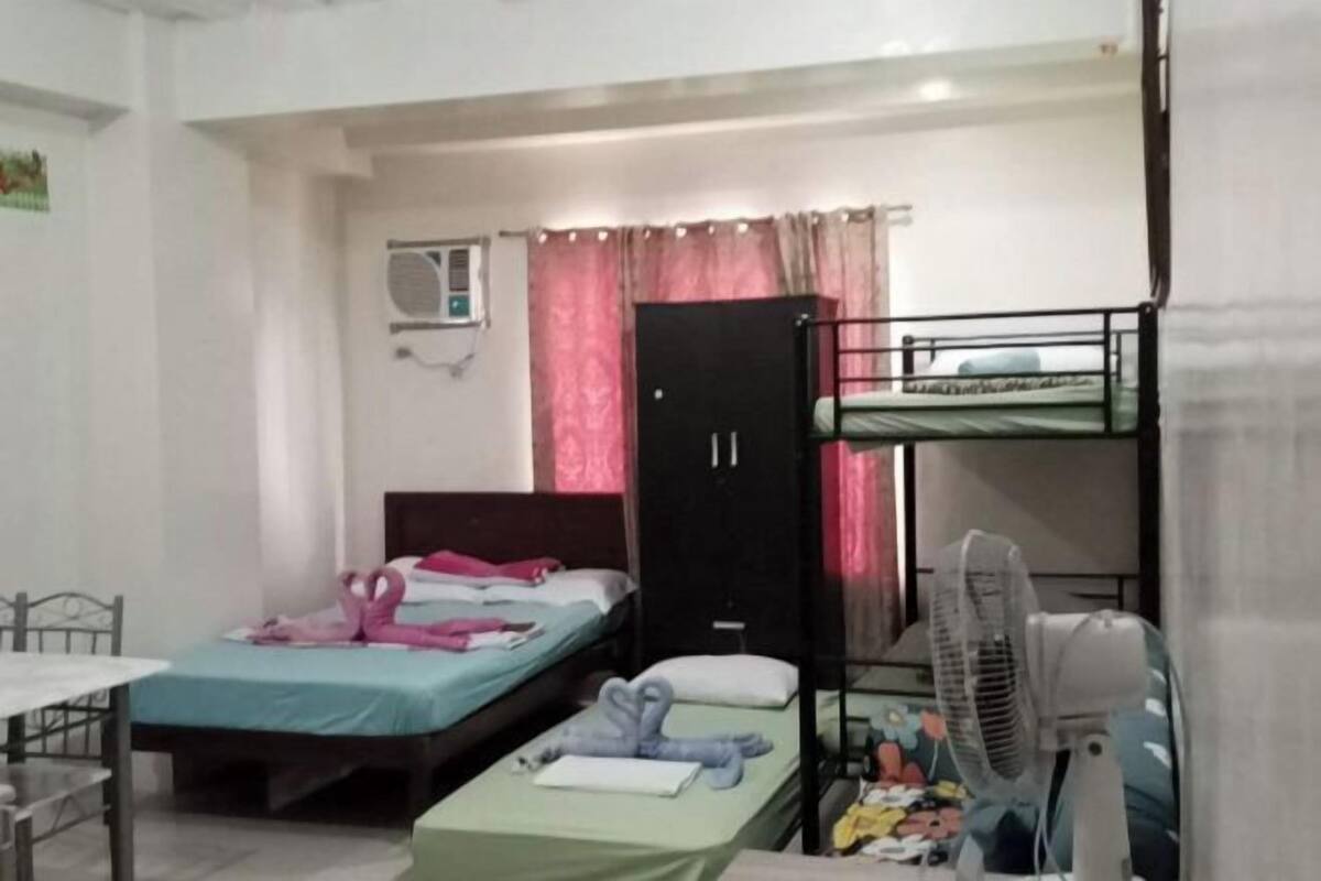 A dormitory room with bunk beds, a desk, a chair, a wardrobe, and a standing fan.