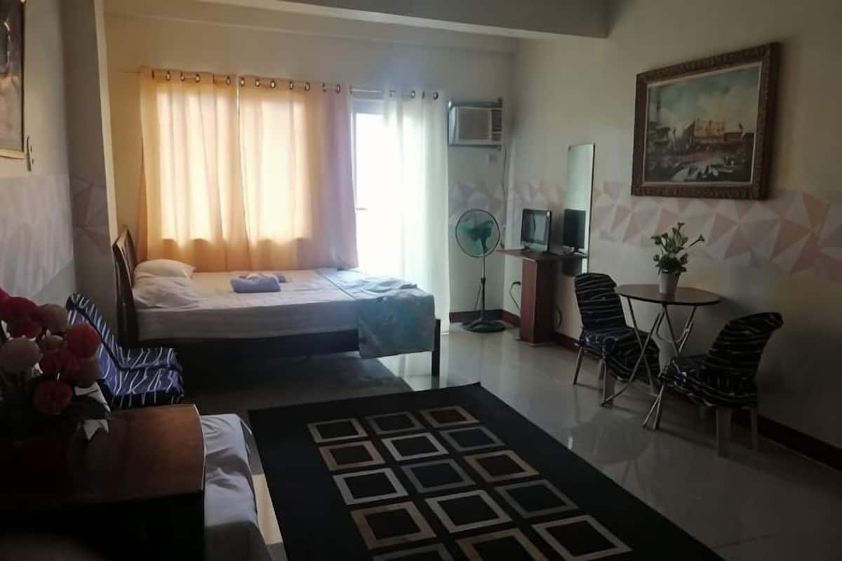A hotel room with a bed, a desk with a chair, a small table with a plant, a standing fan, and a painting on the wall.
