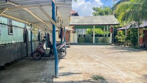 Un espacio exterior techado con una motocicleta estacionada bajo un refugio, un sendero pavimentado y un edificio con techo de tejas al fondo.