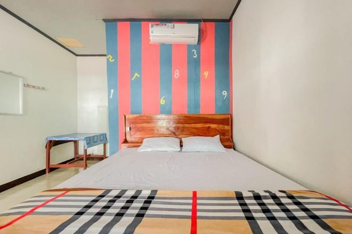 A hotel room with a bed, a wooden headboard, a wall-mounted air conditioner, and a small table with a chair.
