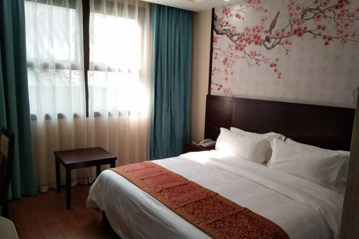 A hotel room with a bed, a small table, a nightstand, and a painting of cherry blossoms on the wall.