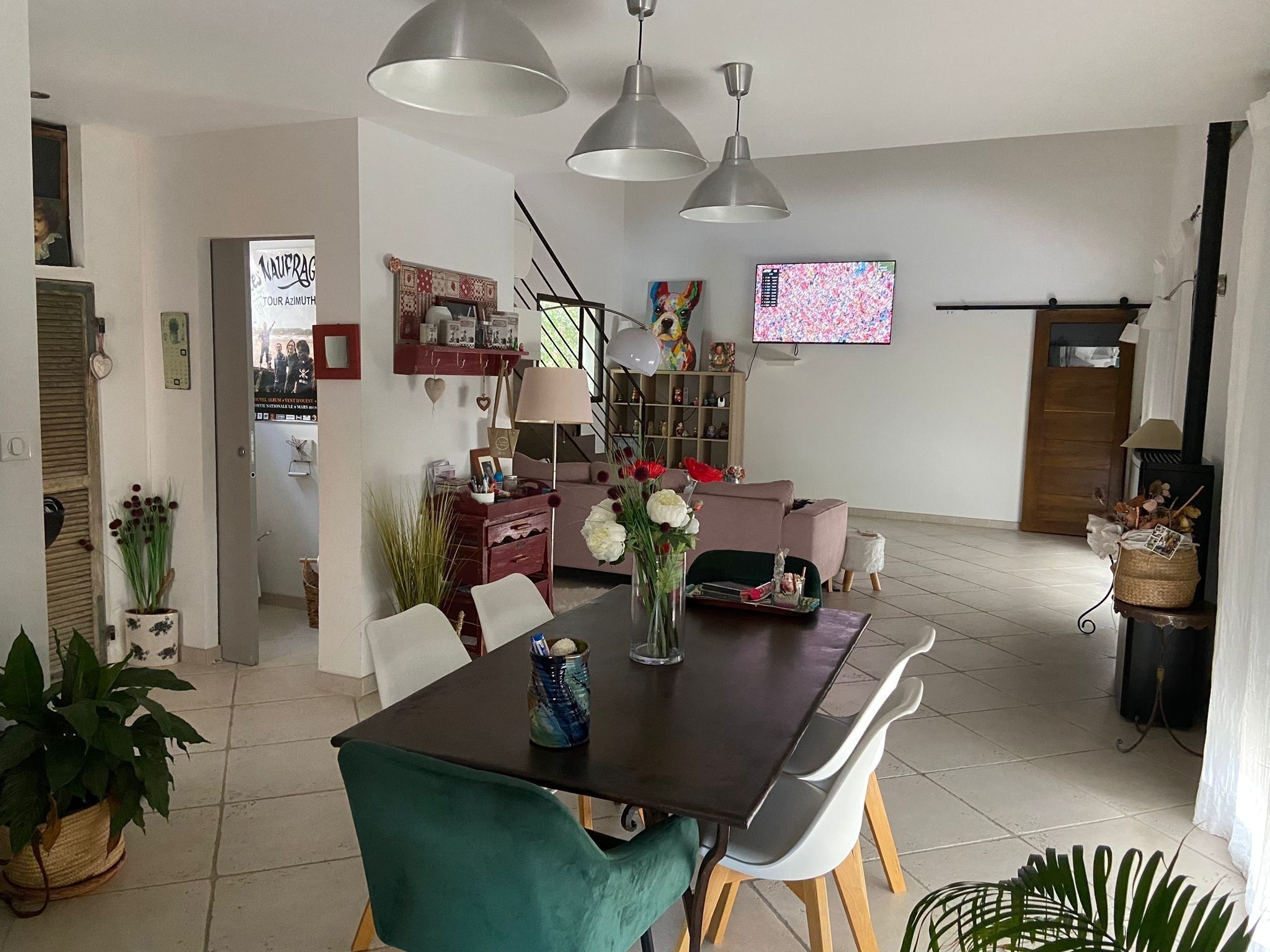 Un coin repas avec une table en bois foncé, des chaises blanches et un fauteuil recouvert d’un tissu turquoise. On y trouve des lampes suspendues, un téléviseur fixé au mur et une bibliothèque contenant divers objets.