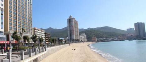 Una playa con un paseo marítimo, un hotel alto y una montaña al fondo.