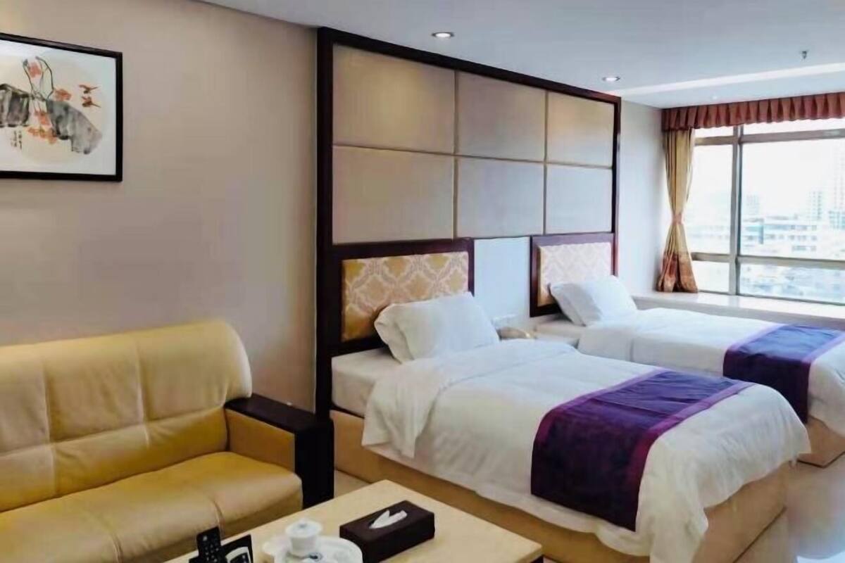 A hotel room with a sofa, two beds, a headboard, a painting, and a window with curtains.