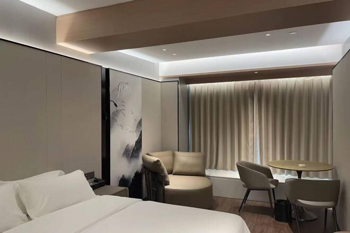 A modern hotel room with a bed, a seating area with chairs, a small table, and a large window with curtains.