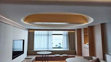 A modern hotel room with a TV mounted on the wall, a seating area with a sofa and two round tables, and a large window with curtains.