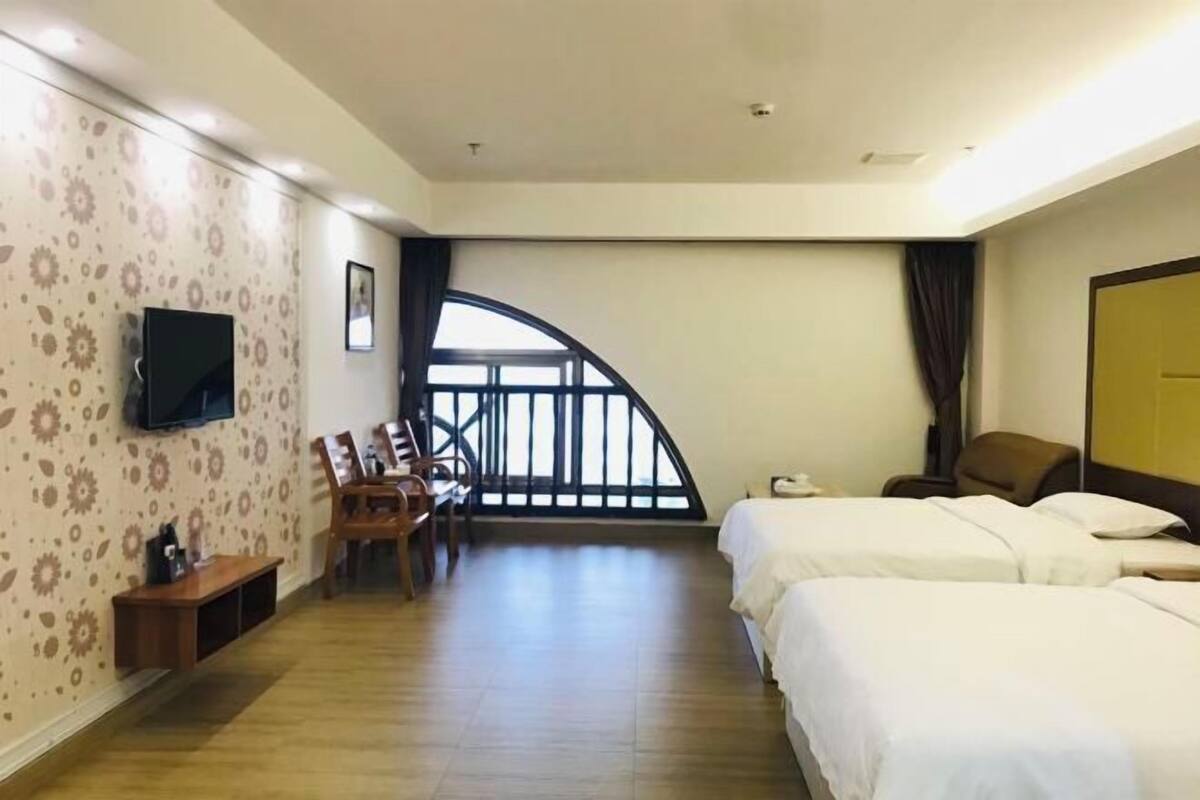 A hotel room with two beds, a TV, a desk with a chair, a wooden bench, and a balcony.