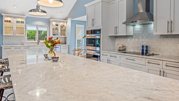 Une cuisine moderne avec des meubles blancs, un grand îlot en marbre, des appareils électroménagers en acier inoxydable et des luminaires suspendus.