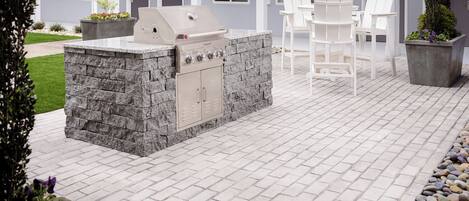 An outdoor patio with a stone grill station, white chairs, and a building with multiple doors in the background.