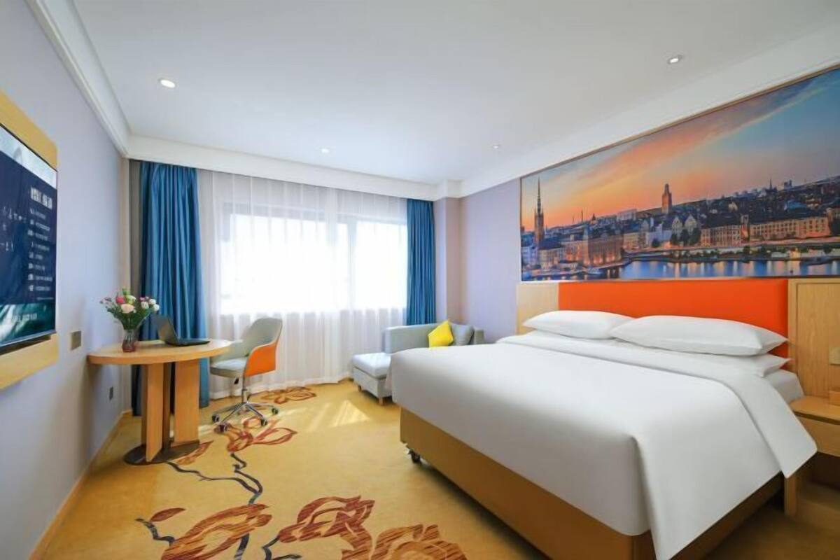 Habitación de hotel con cama, escritorio, silla, ventana con cortinas y un cuadro de paisaje urbano en la pared.