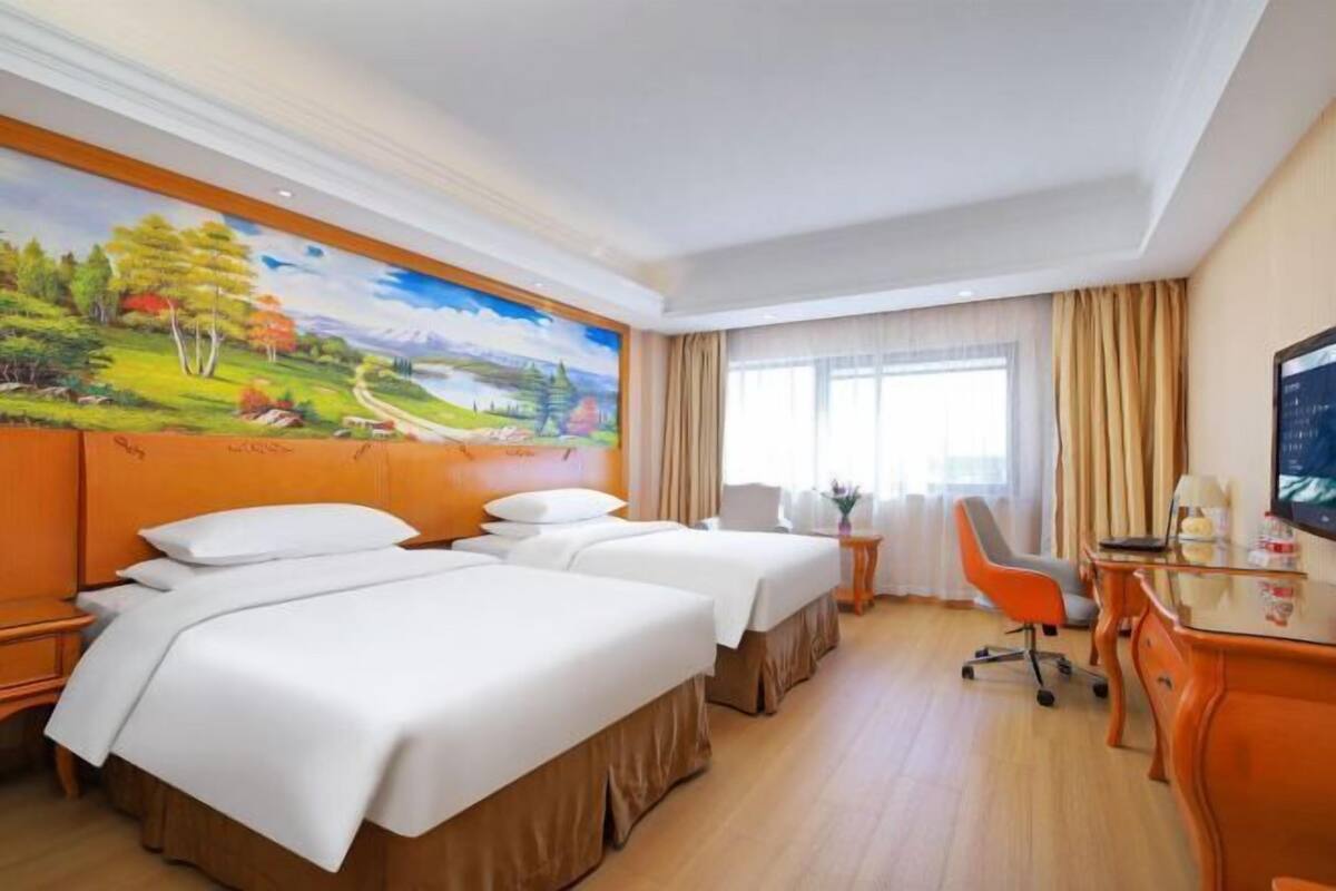 Habitación de hotel con dos camas, un escritorio, una silla, un televisor y un cuadro grande con un paisaje en la pared.