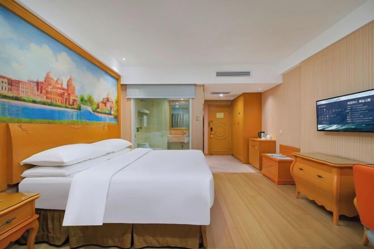Quarto de hotel com cama, escrivaninha, cadeira, televisão e um quadro na parede.