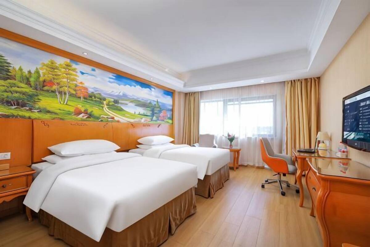 Habitación de hotel con dos camas, un escritorio, una silla, un televisor y un cuadro grande con un paisaje.