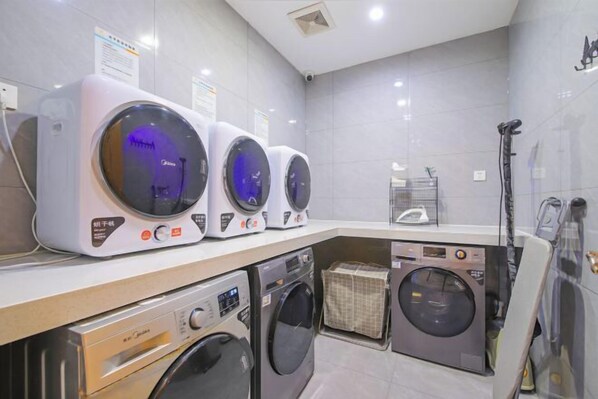 Une buanderie équipée de plusieurs machines à laver et de plusieurs sèche-linge, avec un mur carrelé et une bouche d’aération au plafond.