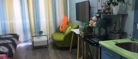A compact living space with a kitchenette, a green chair, a flat-screen TV, and a plant.