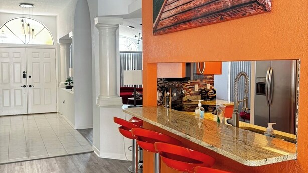 A modern kitchen with a bar area, featuring a wooden bar stool seating, a stainless steel sink, and a large painting of a wooden pier. - Updated luxury 4 Bed W/ KING Bed, FREE Heated Pool, Central Location (Port Charlotte)