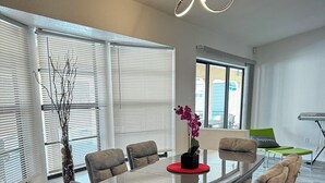 A modern dining room with a glass table surrounded by six grey upholstered chairs, a vase with purple flowers, and a unique circular light fixture. - Updated luxury 4 Bed W/ KING Bed, FREE Heated Pool, Central Location (Port Charlotte)