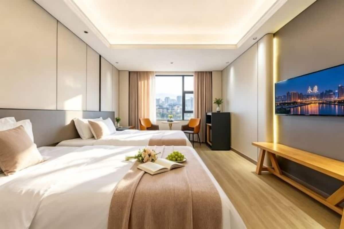 Ein Hotelzimmer mit zwei Betten, einem Schreibtisch, einem Sessel, einem Fernseher und Blick auf die Stadt.